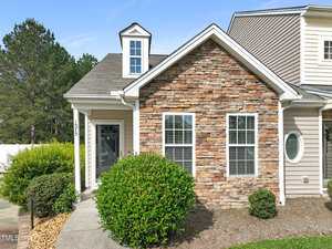 A single-story house with a stone facade, white trim, a dormer window, and a covered entrance. There are bushes and landscaping along the front walkway, and trees are visible in the background.