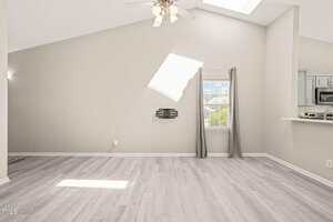 A bright, empty living room with light wood flooring, light gray walls, a ceiling fan, wall-mounted TV bracket, small window with gray curtains, and a partial view of a kitchen with white countertops.