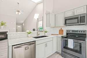 A modern kitchen with light gray cabinets, white countertops, stainless steel dishwasher, black stove with microwave above, white subway tile backsplash, and a sink; part of a living area with a fireplace is visible.