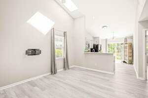 A bright, empty room with light gray wood flooring, light gray walls, a window with gray curtains, a TV wall mount, and an open view into a kitchen with stainless appliances and a sunlit dining area.