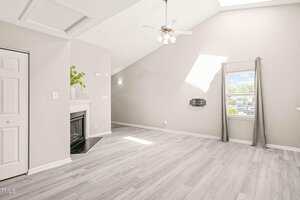 A spacious, empty living room with light-colored wood flooring, a corner fireplace with a plant on the mantle, a ceiling fan, a window with gray curtains, and sunlight streaming through a skylight.