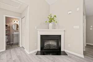 A white fireplace with a black hearth sits against a beige wall. A large white vase with green branches decorates the mantel. To the left, an open door reveals a bathroom with a round mirror and vanity.