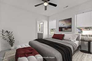 A bedroom with a large bed featuring gray and red bedding, a small side table with a lamp, a framed artwork on the wall, two windows, a ceiling fan, a plant in the corner, and an open doorway to a bathroom.