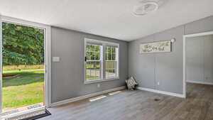 A bright room with light wood flooring, gray walls, two large windows, an open door showing green grass and trees outside, and a decorative sign on the wall reading "bless our nest.