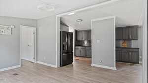 A modern kitchen with dark cabinets, a black refrigerator, light wood floors, and light gray walls. A doorway leads to another room, and there is a decorative sign on one wall.