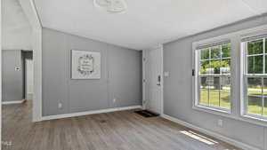 A bright, unfurnished room with light wood floors, gray walls, a white entry door, large windows letting in sunlight, and a framed sign on the wall that reads "Love makes a house a home.