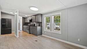 A modern kitchen with dark cabinets, stainless steel appliances, and light wood flooring, adjacent to a bright living area with large windows overlooking a driveway with parked cars.