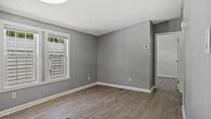 A small, empty room with gray walls, light wood flooring, two windows with white trim, and an open doorway leading to another area with similar flooring.