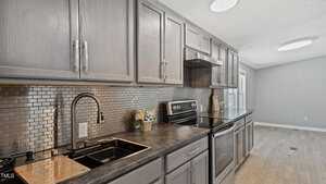 Galley kitchen with gray cabinets, dark countertops, stainless steel backsplash tiles, a black sink with a gooseneck faucet, an electric range, and light wood floors. Natural light enters from a window at the far end.