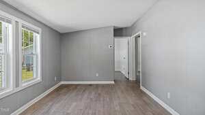 A bright, empty room with light gray walls, wood-style flooring, and three large windows on the left. The room leads to a hallway with white doors and has a white ceiling.