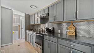 Modern kitchen with gray cabinets, stainless steel appliances, tile backsplash, and light wood flooring. Countertops are dark, and a decorative cutting board leans against the wall near the stove. Doorways are visible in the background.