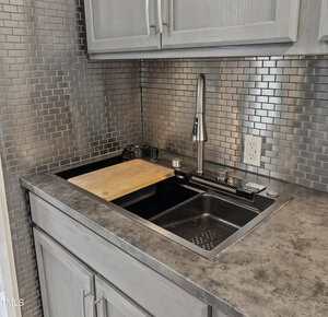 A modern kitchen sink with a black basin, stainless steel faucet, gray cabinets, metallic tile backsplash, and a cutting board partially covering the sink. There is an electrical outlet on the backsplash.
