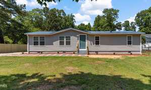 A single-story house with gray siding, a blue metal roof, and a central front door accessed by steps. The yard is grassy with a stone walkway and small shrubs along the front of the house. Trees are in the background.