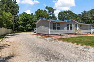 Single-story manufactured home with light gray siding, blue roof trim, and multiple windows. A small wooden staircase leads to the entrance. Gravel driveway and green lawn with trees in the background.