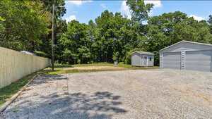 A gravel driveway leads to a metal garage and a small shed, both situated near a grassy area and surrounded by trees. A wooden fence runs along the left side of the property under a clear sky.