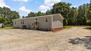 A single-story manufactured home with light gray siding, white trim, and a front porch with steps, sits on a gravel lot surrounded by trees and grass under a partly cloudy sky.