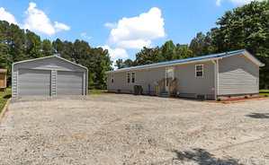 Single-story gray manufactured home with a metal roof, adjacent to a detached gray metal garage, situated on a gravel driveway with trees and a lawn in the background under a partly cloudy sky.