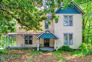 The two-story, cream-colored house at 903 Nicholas Street in Henderson features peeling paint and a covered front porch supported by four posts. Large tree branches frame the yard, where scattered leaves and overgrown grass add rustic charm.