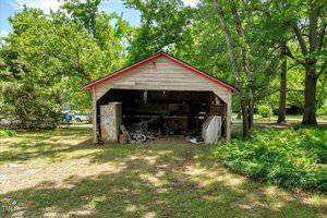 A weathered, open-front shed in a grassy yard with trees, containing assorted items including an old metal cabinet, scrap materials, and bicycles. The shed has a red roof and beige siding.