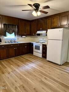 A kitchen with dark wooden cabinets, light wood flooring, white appliances including a refrigerator and stove, a ceiling fan with lights, and a small window above the sink with curtains.