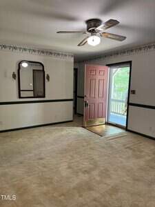 A room with beige carpet, a ceiling fan with a light, a door painted red standing open to a porch, white walls with green trim, wallpaper border, and a wall mirror with two sconces.