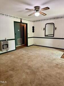 A carpeted room with white walls, green trim, a ceiling fan with a light, a wall mirror, a wall-mounted heater, and floral border near the ceiling. An open doorway leads to another area with wood flooring.