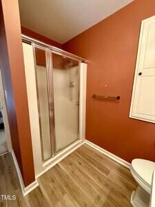 A bathroom with reddish-brown walls, a glass shower enclosure with a sliding door, a towel bar, a white wall cabinet, wood-look flooring, and a white toilet partially visible in the lower right corner.