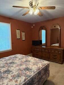 A bedroom with rust-colored walls, a ceiling fan with lights, a large wooden dresser with two mirrors, a small TV, a bed with a floral-patterned mattress, and two framed pictures on the wall next to a window.
