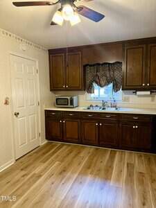 A kitchen with wood cabinets, white countertops, a stainless steel sink under a window with a dark valance, a microwave on the counter, light wood flooring, a ceiling fan with lights, and a closed white door.
