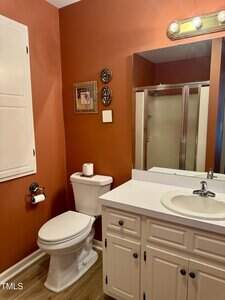 A bathroom with rust-colored walls, a white toilet, wall-mounted toilet paper holder, white vanity with sink, large mirror, and a shower with a glass door. There is framed wall decor above the toilet.