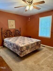 A bedroom with tan carpet, orange-brown walls, a ceiling fan with lights, a window with blinds, a wooden bed frame, and a floral-patterned mattress. A framed picture hangs above the bed.