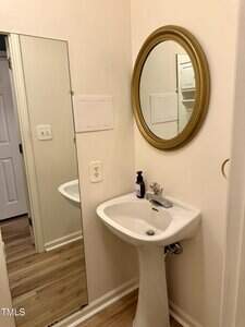 A small bathroom corner with a white pedestal sink, a soap dispenser, a gold-framed round mirror above the sink, and a tall rectangular mirror on the adjacent wall. Light wood flooring and neutral walls are visible.