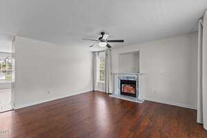 A room with wood flooring, a white ceiling fan, a window with curtains, and a fireplace with a marble surround and a small alcove above it. The walls are light-colored and the space is unfurnished.