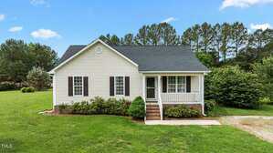A single-story white house with black shutters at 3803 Ridgewood Drive, Franklinton, features a small covered front porch and brick steps, surrounded by green grass and trees under a clear blue sky.