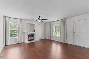 A living room with hardwood floors, a ceiling fan, a white fireplace with a marble surround, large windows with curtains, and a white front door. The walls are painted light gray.