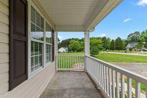 A covered front porch with white railings, beige siding, and a window with brown shutters overlooks a green lawn and a suburban street with trees and neighboring houses.