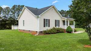 A single-story beige house with white trim and dark shutters, small front porch, and brick foundation sits on a large green lawn with trees in the background and a white shed visible to the left.