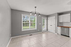 A bright, unfurnished kitchen area with beige tile flooring, gray walls, white trim, and large windows with white blinds. There is a glass light fixture, a door, and some white cabinetry with a wooden countertop.