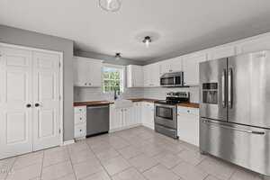 Modern kitchen with white cabinets, stainless steel appliances, wood countertops, a farmhouse sink under a window, and light gray tile flooring. A double-door pantry is on the left side of the image.