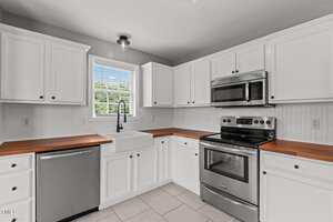 A modern kitchen with white cabinets, wood countertops, stainless steel appliances including a stove, microwave, and dishwasher, a farmhouse sink under a window, and light gray walls.