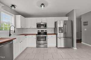 Modern kitchen with white cabinets, stainless steel appliances including a refrigerator, oven, microwave, and dishwasher, wood countertops, tile floor, large window with blinds, and gray walls.