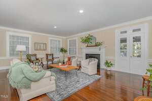Bright living room with hardwood floors, a beige sofa, two armchairs, a gray fireplace, white trim, a large rug, houseplants, and French doors leading outside. Multiple windows let in natural light.