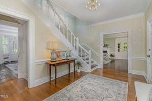 A bright entryway with light-colored walls, hardwood floors, a patterned rug, and a wooden console table with a lamp, framed art, and plants. A staircase leads upward, and doorways open to other rooms.