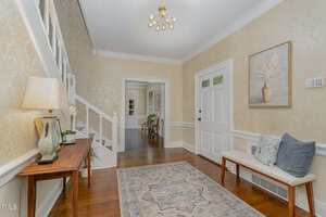 A bright hallway with light yellow patterned walls, a white front door, a wooden bench with cushions, a console table with a lamp and flowers, a rug, and a staircase with white railing. A modern ceiling light is overhead.