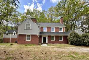 Charming two-story brick and wood-sided house at 938 Hargrove Street in Henderson, with a gabled roof, multiple chimneys, white-trimmed windows, and a patchy front yard nestled among trees under a partly cloudy sky.