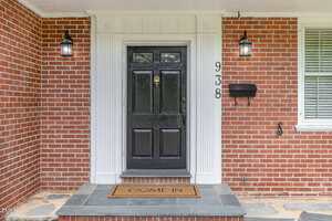 A black front door with a brass knocker and white trim is centered on a red brick house. There are two black lantern-style lights, a mailbox, house numbers "938", and a doormat that says "COME IN.