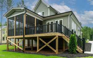 A gray house with white trim features a raised wooden deck with black railings and a screened-in porch area. The yard is landscaped with mulch and green grass, and trees are visible in the background.