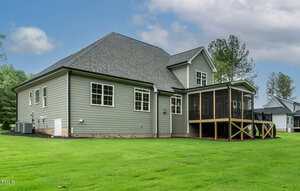 A two-story house with gray siding, white trim, and a screened-in porch. The yard is green and well-maintained, and mature trees and neighboring houses are visible in the background under a partly cloudy sky.