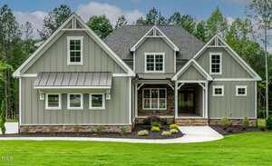 A modern two-story house with gray board-and-batten siding, white trim, stone accents, and large windows, set on a manicured lawn with young landscaping and surrounded by trees.