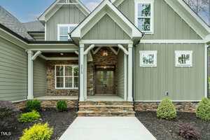 A front view of a modern house with green vertical siding, stone accents, white trim, a covered entryway with wooden double doors, and landscaped shrubs along the walkway.